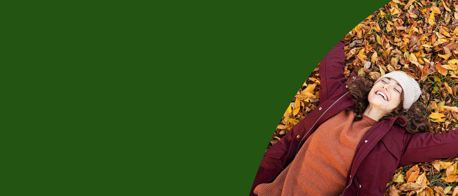 A woman lying on a bed of autumn leaves, wearing a burgundy jacket, rust-colored sweater, and white beanie, set against a solid dark green background.- desktop