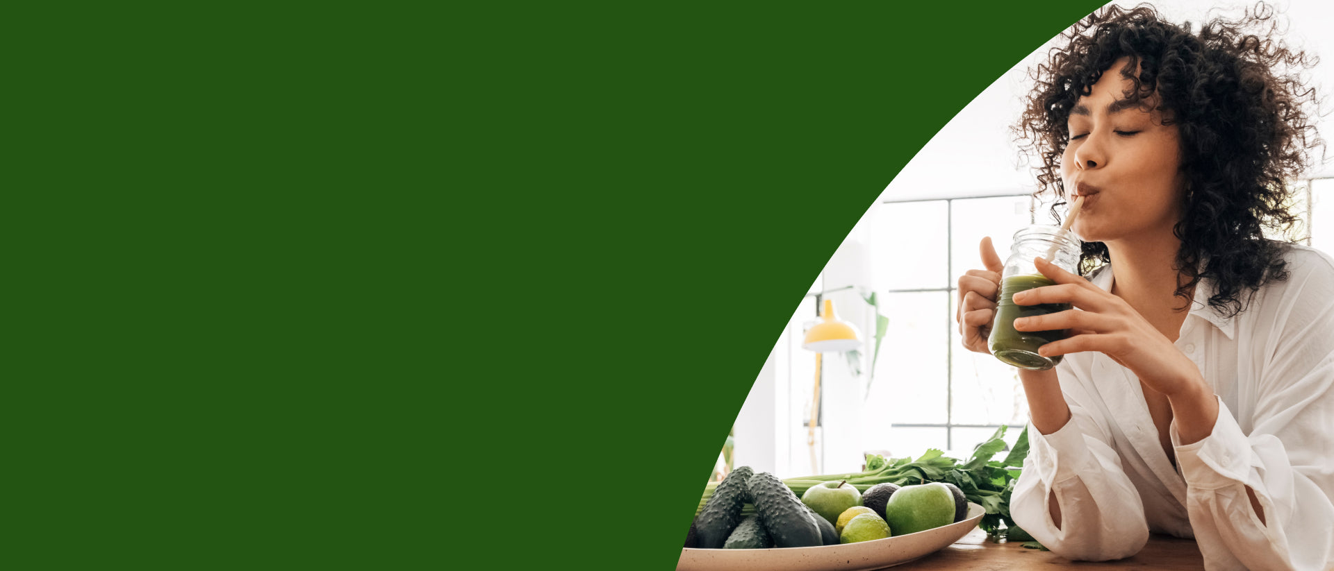A woman with curly dark hair wearing a white blouse is drinking a green smoothie from a glass jar using a straw. She is sitting at a wooden table with fresh vegetables, including cucumbers, limes, and greens, in a bright kitchen setting.- desktop