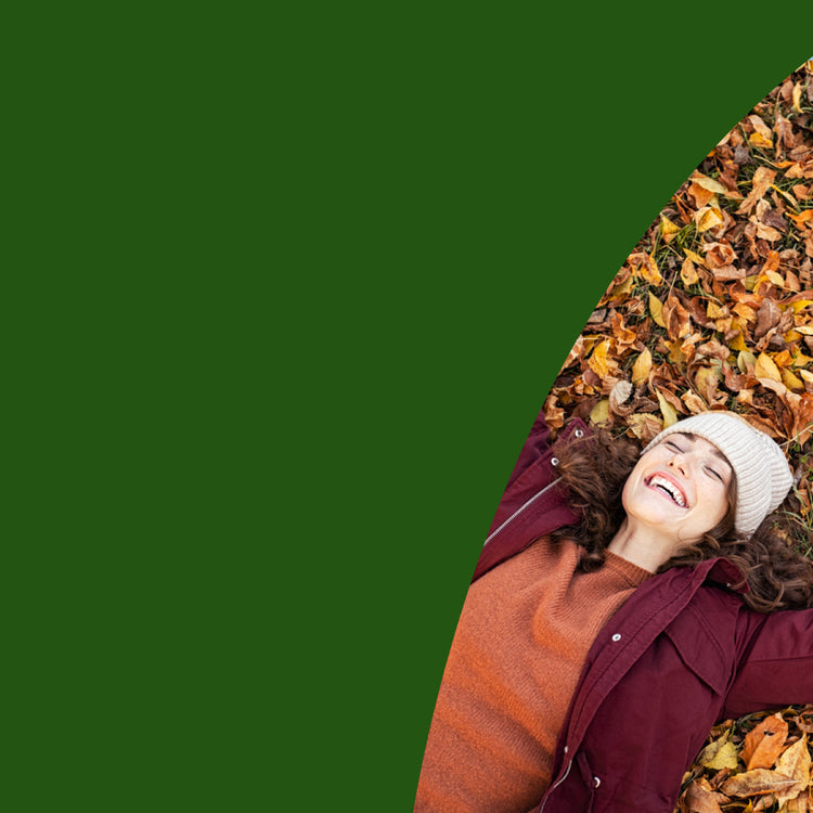 A woman lying on her back on a bed of autumn leaves, wearing a burgundy jacket, rust-colored sweater, and white knit beanie, set against a solid dark green background. - mobile