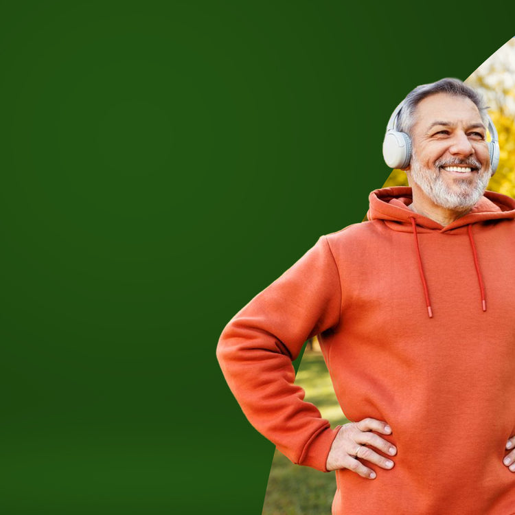 A smiling older man with gray hair and a beard, wearing white over-ear headphones and an orange-red hoodie, standing outdoors against a solid green background with a partial diagonal cutout of a park scene.- mobile