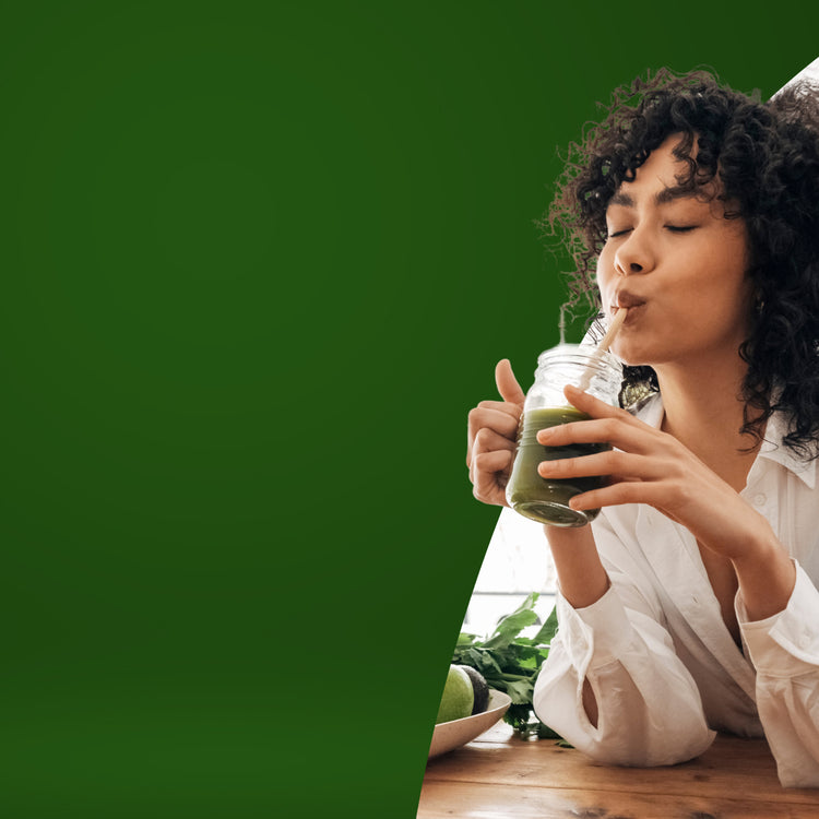 A woman with curly dark hair wearing a white button-down shirt drinks a green smoothie from a glass jar with a metal straw. She is seated at a wooden table with green leaves and an apple nearby, set against a solid dark green background. - mobile