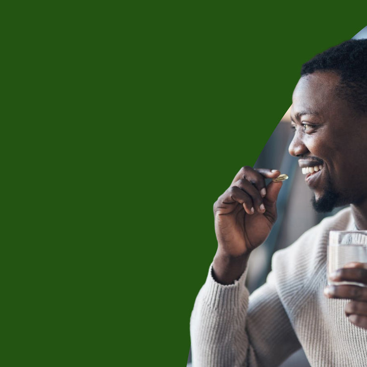 A smiling man in a light beige knit sweater holds a small gold capsule near his mouth while holding a glass of water, set against a solid dark green background.- mobile